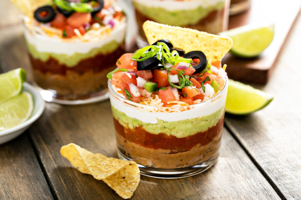 Mini layered taco dip cups on a wooden surface with chips and lime
