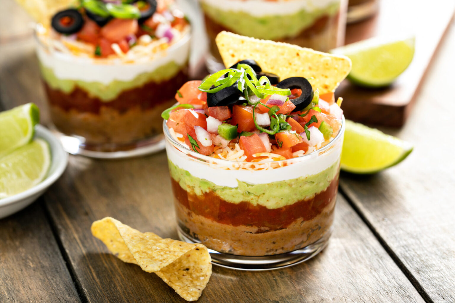 Mini layered taco dip cups on a wooden surface with chips and lime