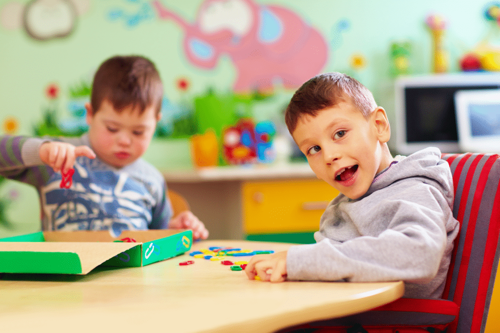 a little boy with cerebal palsy sits at a table. One looks happy and directly at the camera, the other is in the background