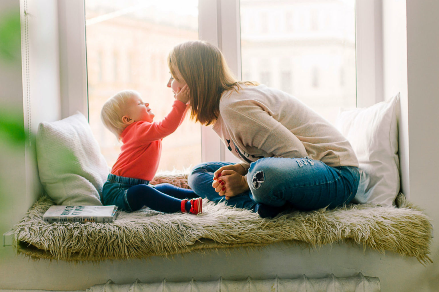 toddler reaching for mother's face as they sit facing each other in a windowsill