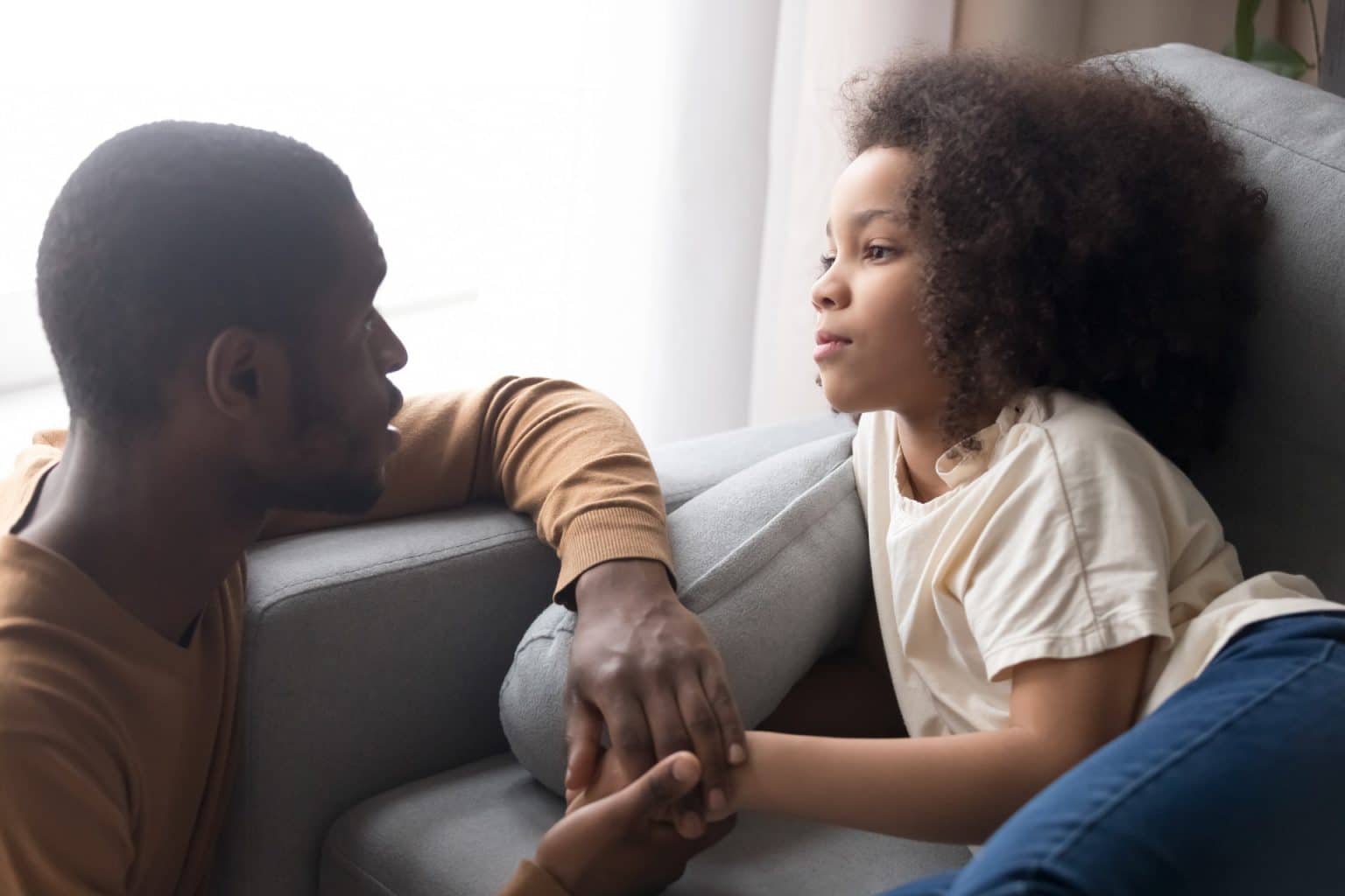 Father and daughter on the couch having a talk. The father looks compassionate and the daughter looks thoughtful