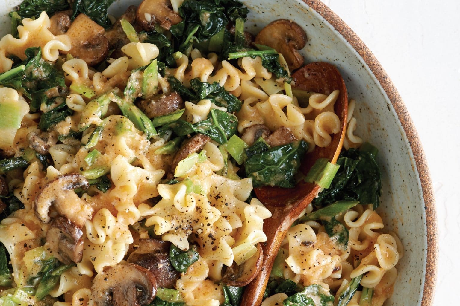 bowl of pasta with greens and mushrooms. A wooden spoon rests in the bowl