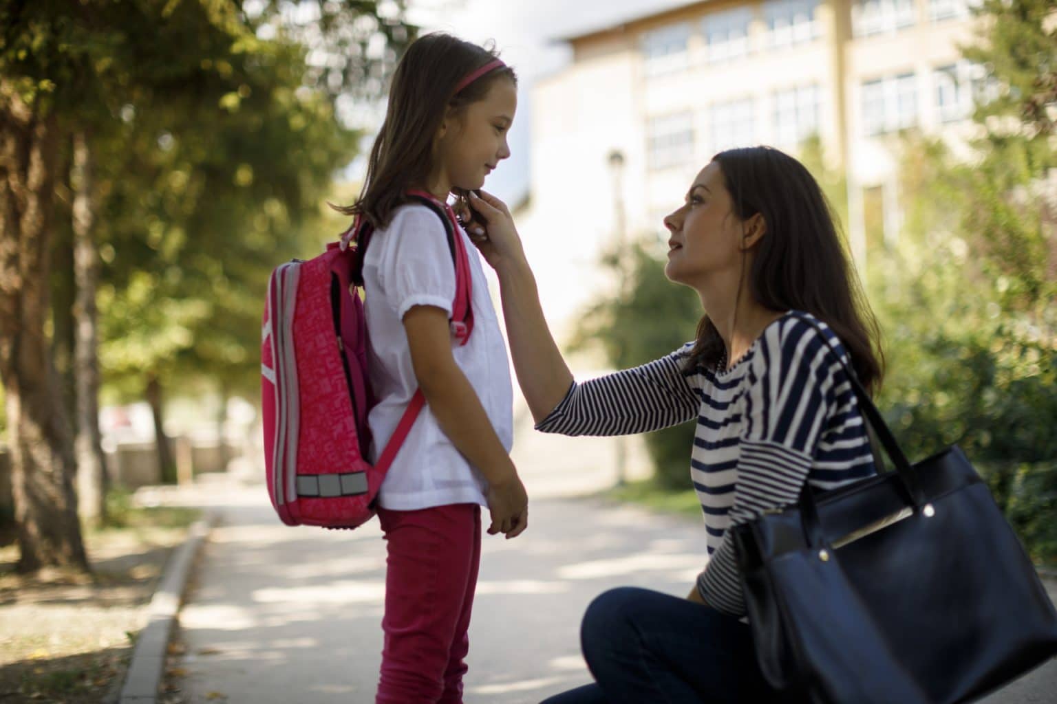 mother bends down to hold daughter's face. Daughter is wearing a backpack as if ready to go to school