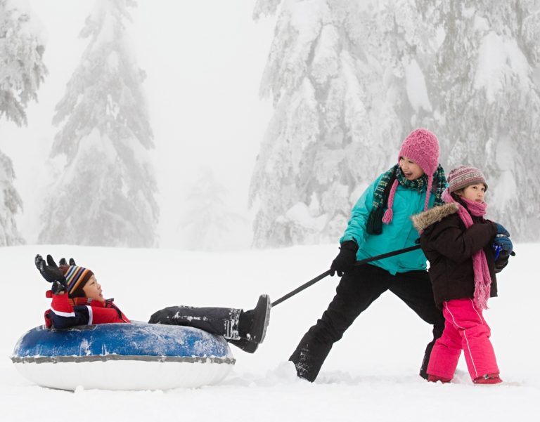 Three kids outside on a snowy day, one is on a blue sled being pulled by the other two by a long rope.