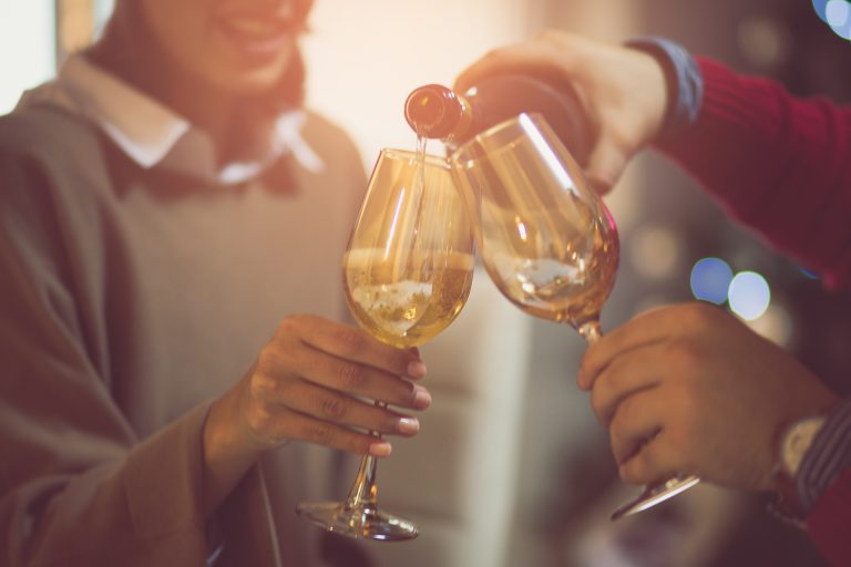 Two adults maybe parents drinking alcohol together, close-up of wine glasses as arm of one adult pours more wine. Cropped face of female-presenting person smiling in blue sweater.