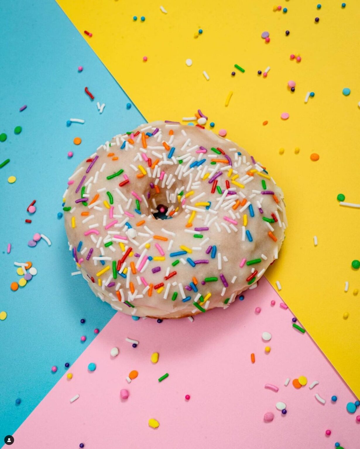 A donut with rainbow sprinkles on a blue, yellow, and pink background from a hidden gem in Toronto, Glory Hole Donuts.