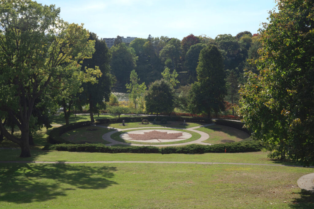 A Toronto park, High Park, on a sunny day. Large trees surround a large decorative maple leaf in the centre of a clearing.