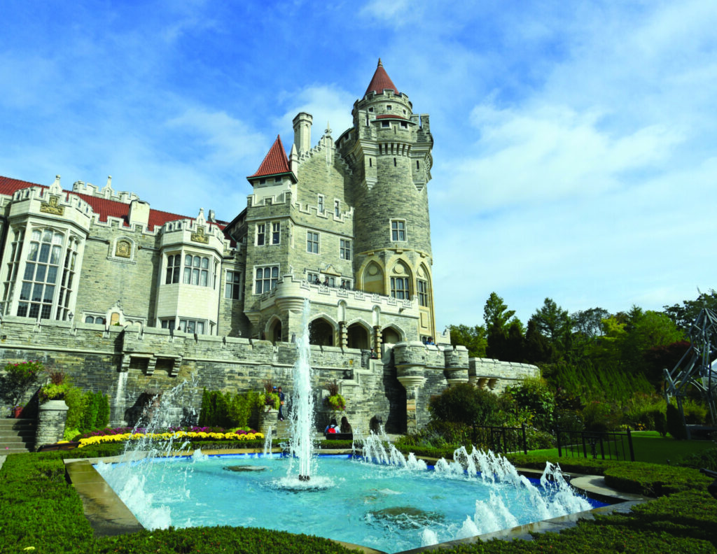 Casa Loma, a large castle structure in the middle of Toronto, with a blue fountain, a bed of flowers, and trees in front of it.