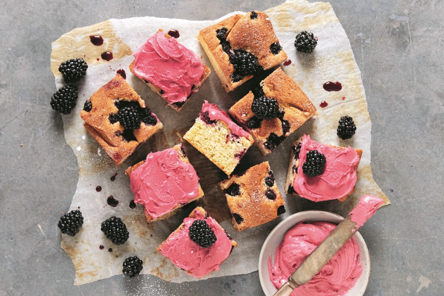 Squares of blackberry almond snacking cake with bright pink berry icing sit on parchment paper