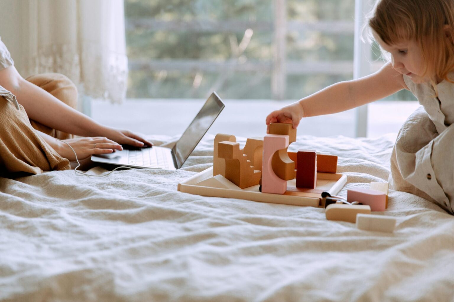 Woman working on computer in front of child playing with blocks, to illustrate intensive mothering