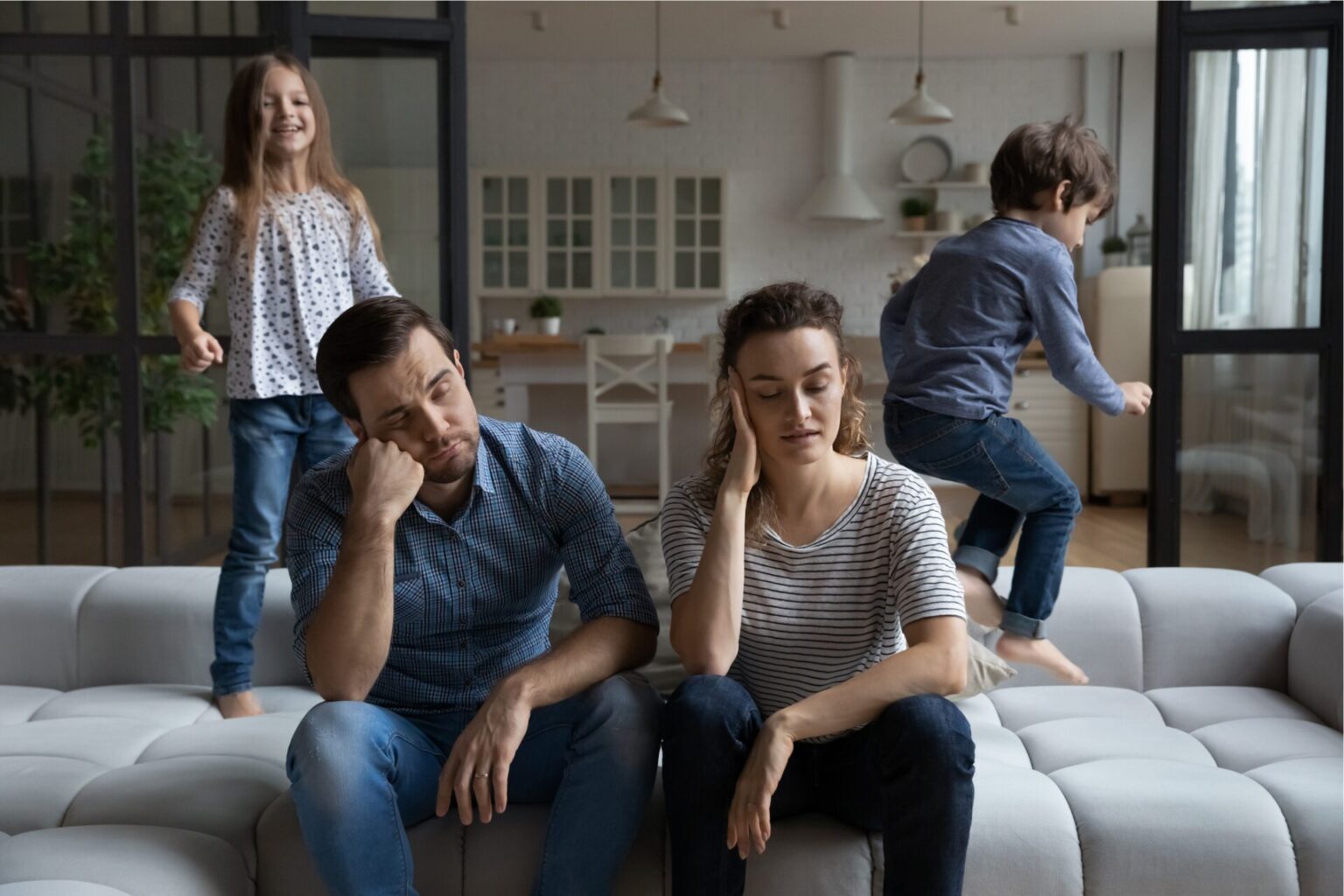 Two parents sit side-by-side looking frustrated as their kids play behind them