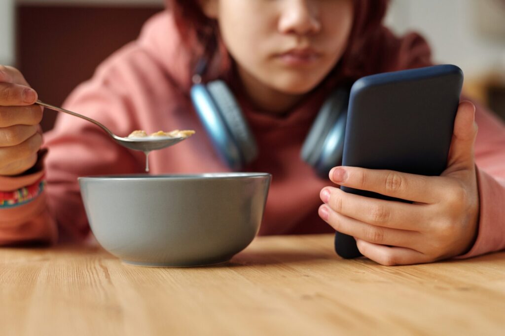 A kid eats breakfast while holding their phone, sucked in by screen time
