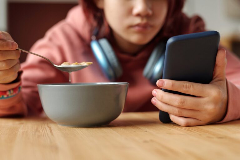 A kid eats breakfast while holding their phone, sucked in by screen time