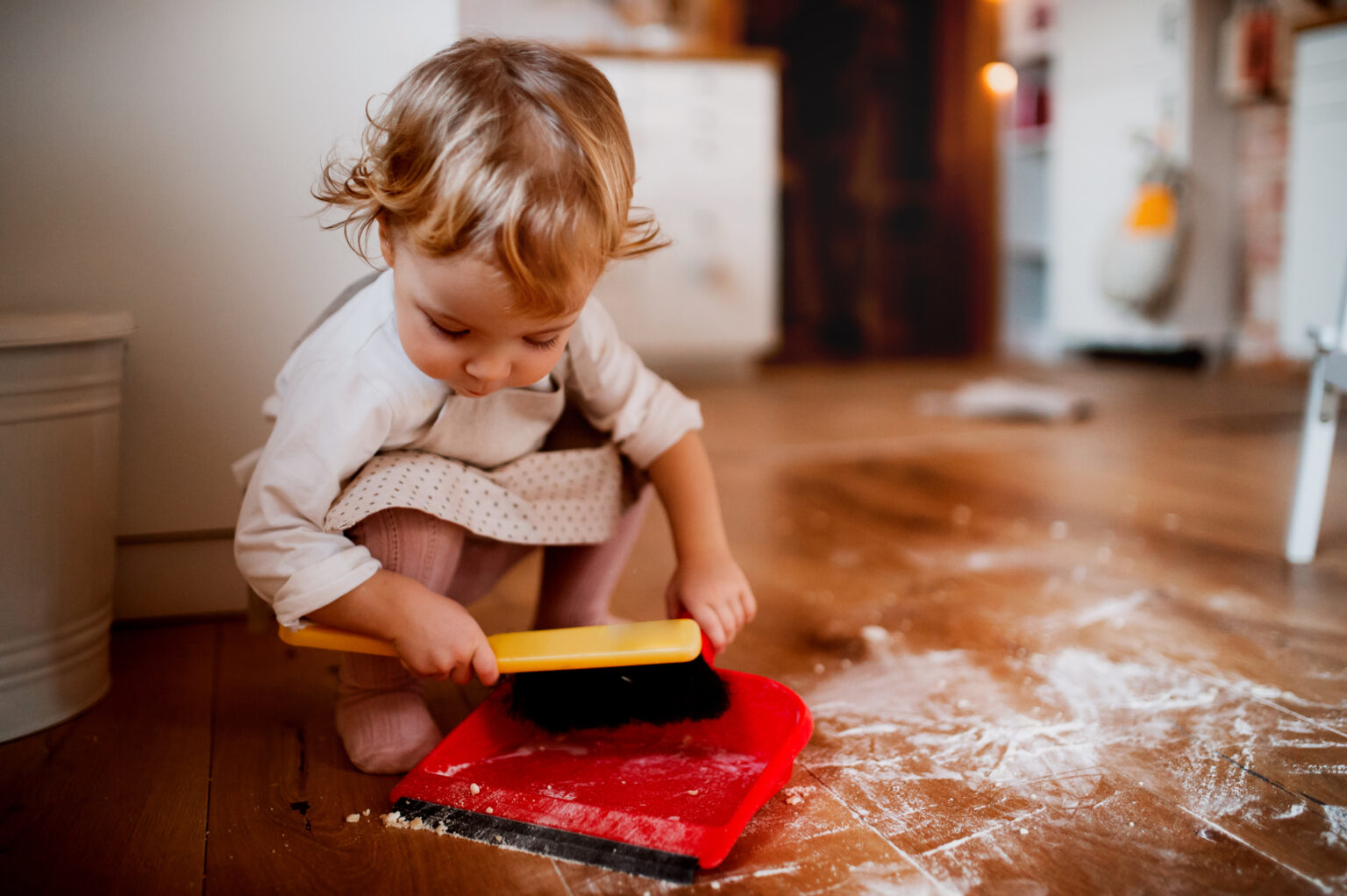 A toddler helping to sweep into a red dustpan, to illustrate ways to let toddlers help